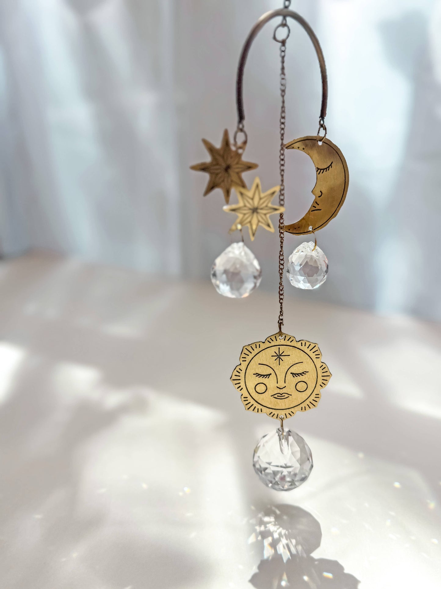 A celestial gold-toned metal suncatcher ornament with a crescent moon, stars, and a sun face charm, accented by clear faceted crystal teardrop pendants, hanging against a softly lit white background.