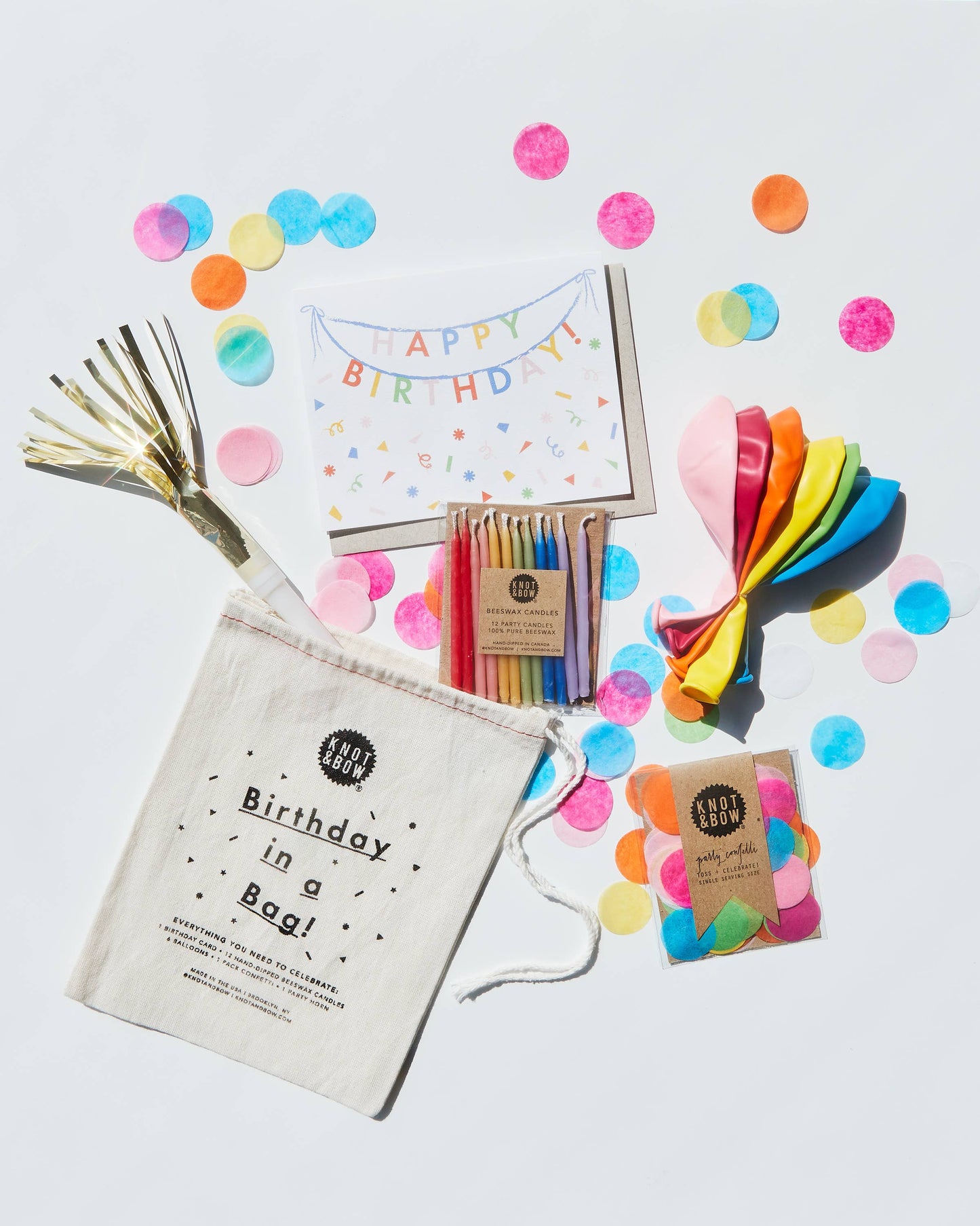 Birthday party supplies including candles, balloons, and a bag labeled 'Birthday in a Bag' on a light gray background with confetti.