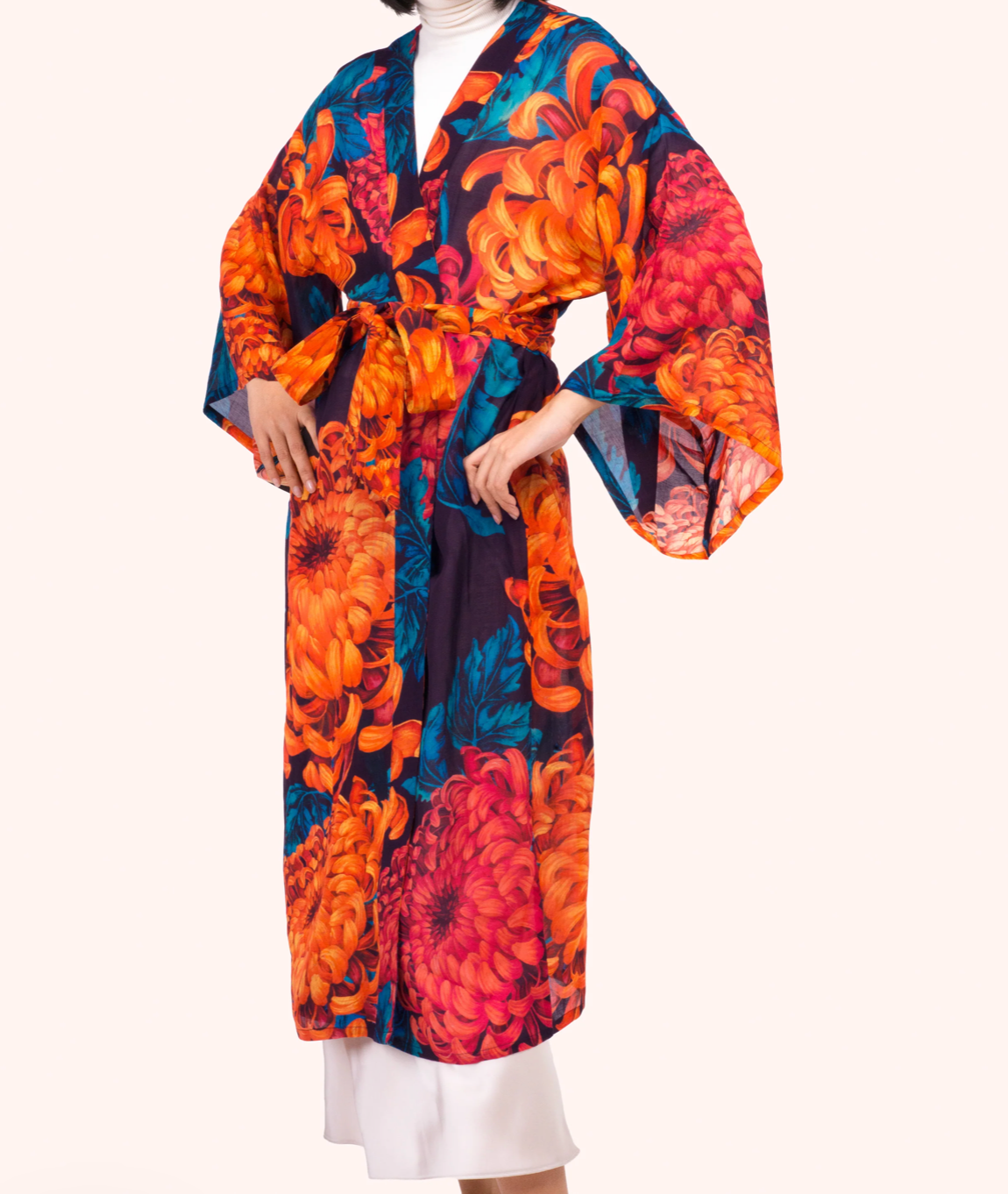 A model wearing a long-sleeved kimono-style robe with a bold floral print in orange, red, and teal on a dark background, styled over a white turtleneck and white skirt against a solid cream backdrop.