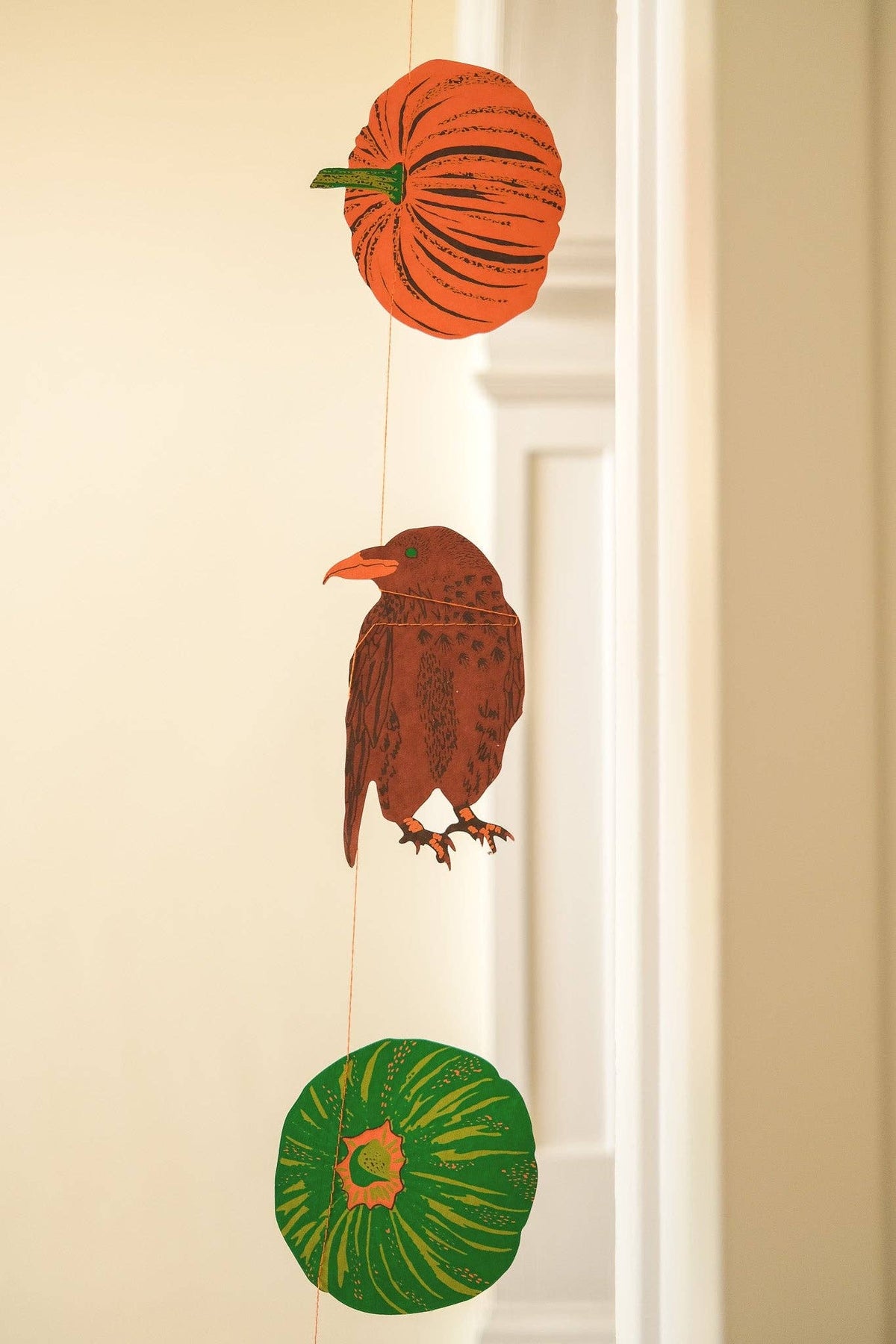 A hanging autumn decoration featuring three illustrated paper-cut style shapes: an orange pumpkin at the top, a brown bird in the middle, and a green pumpkin at the bottom, suspended by thin strings against a warm beige interior wall.
