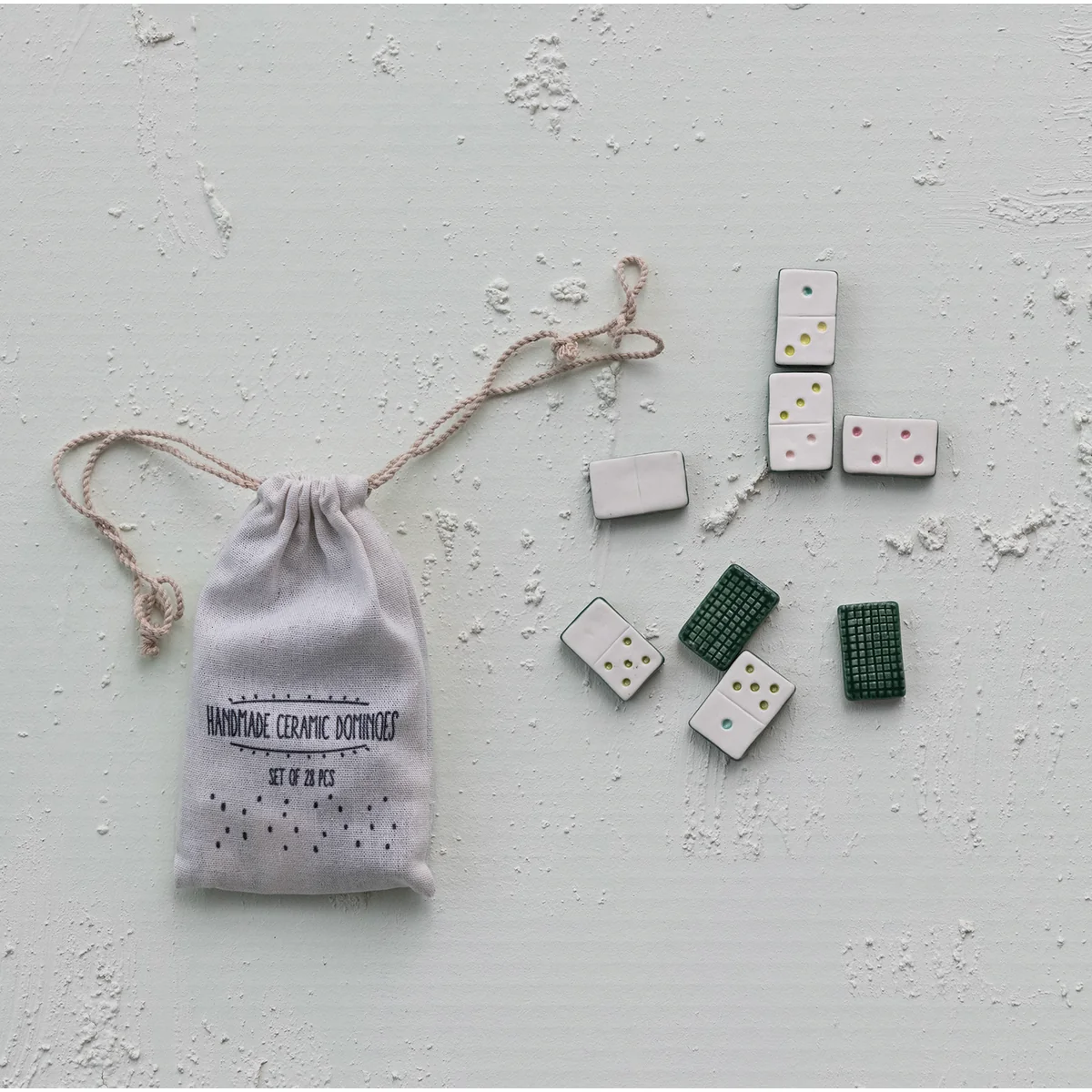 A set of ceramic dominoes with white, green, and dark gray tiles, displayed next to a small white drawstring pouch printed with 'HANDMADE CERAMIC DOMINOES' on a textured light gray background.