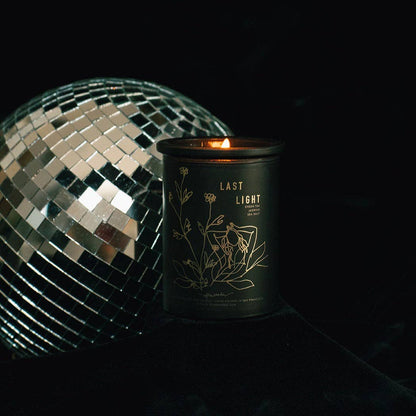 A black candle tin with gold botanical line art and the text 'LAST LIGHT' sits lit on a dark surface beside a mirrored disco ball against a black background.