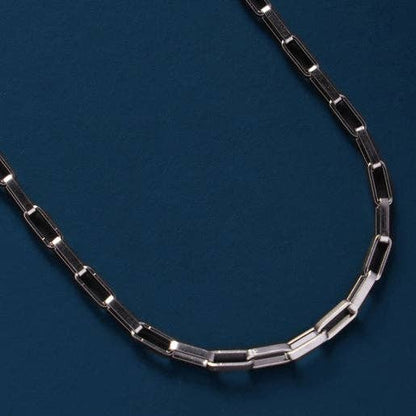 A silver-toned paperclip chain necklace displayed in a curved shape against a dark blue textured background.