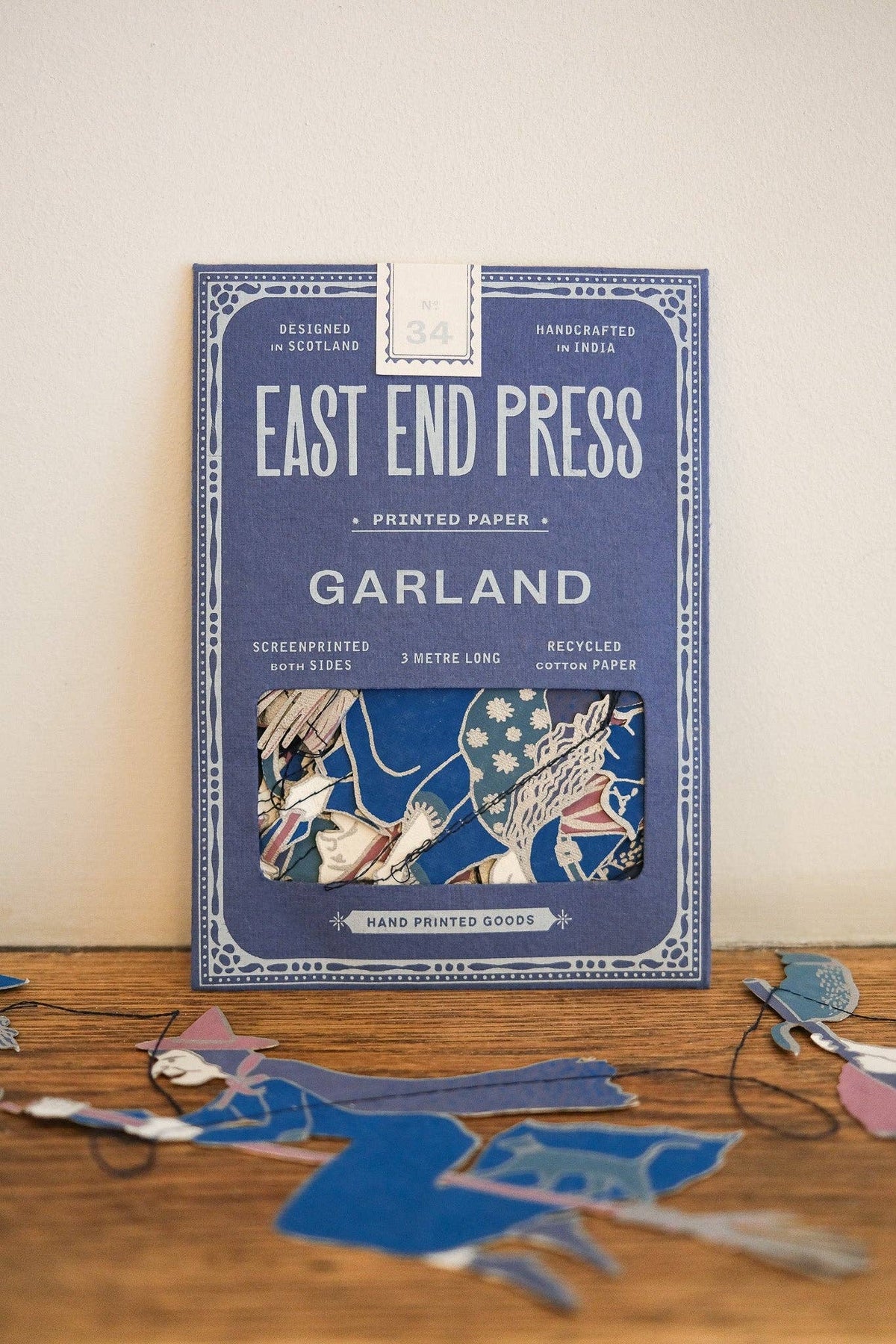 A package of East End Press Garland printed paper stands upright on a wooden surface against a light beige wall. The box is blue with white text and decorative border details, and a small window near the bottom shows the layered paper artwork.
