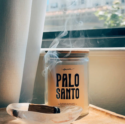 A glass jar candle labeled 'PALO SANTO' sits on a wooden surface near a window with smoke rising from the top. A smudge rests in a clear resin dish in the foreground.
