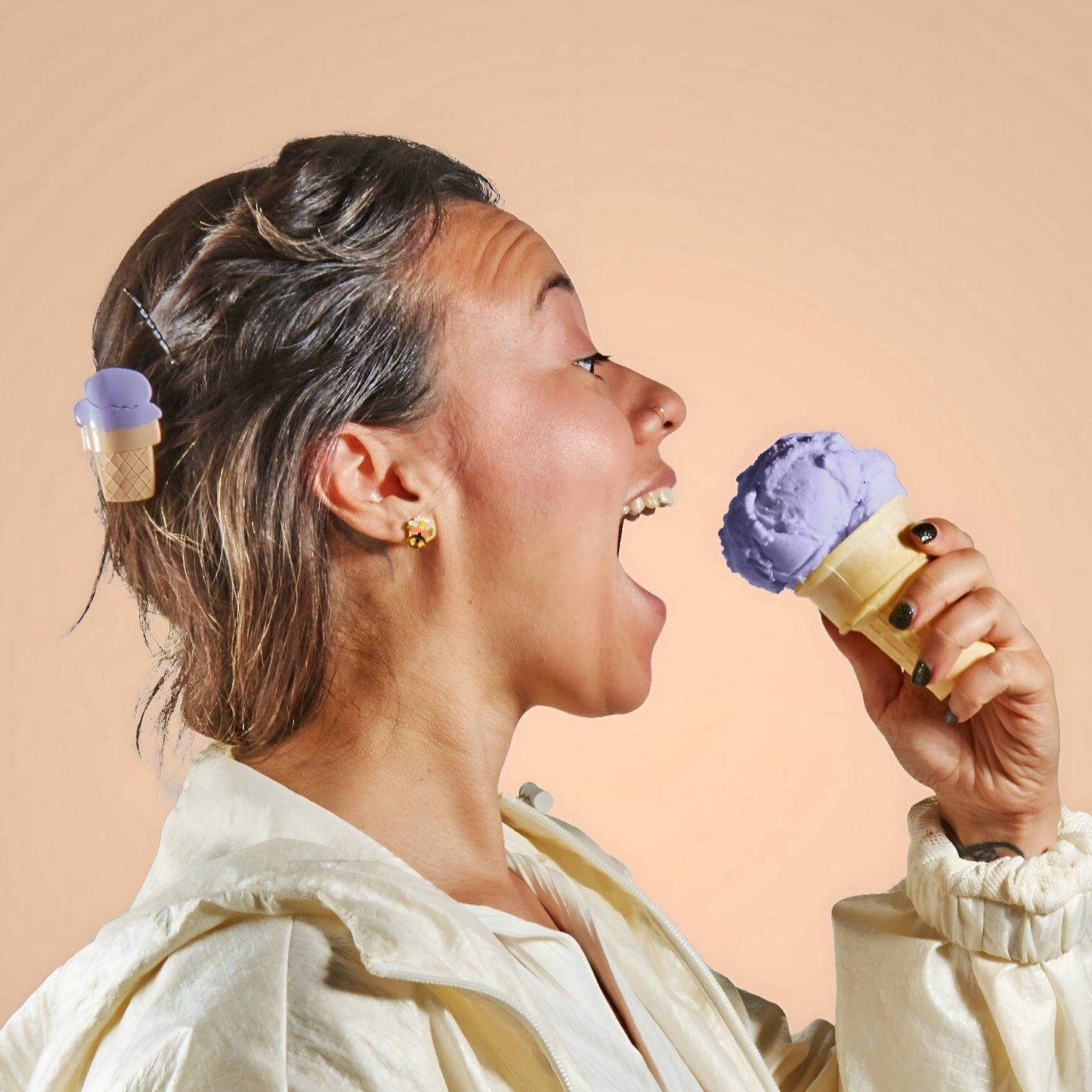 Person eating a purple ice cream cone with another ice cream cone in their hair against a beige background