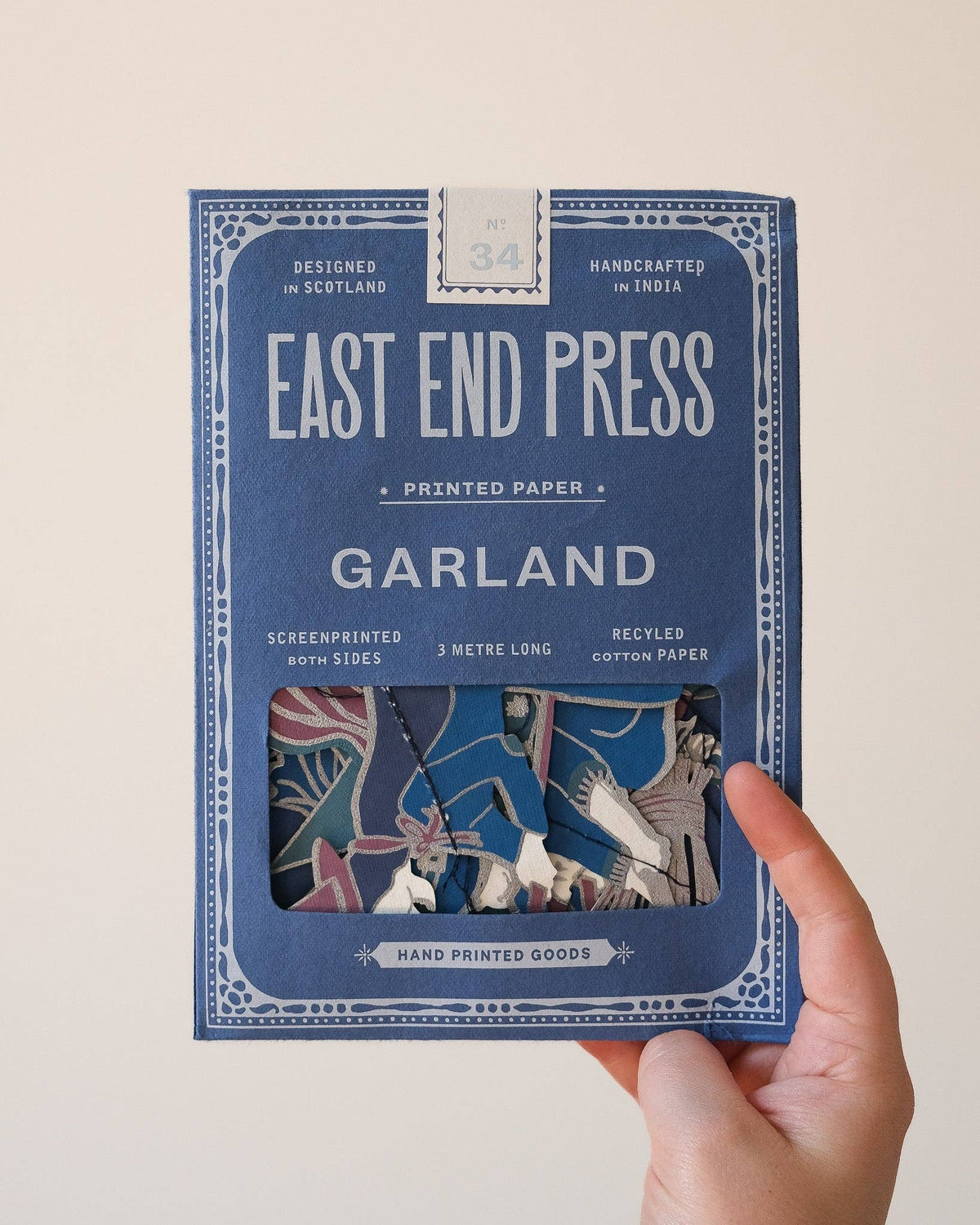 A hand holding a blue package of East End Press Garland printed paper against a plain light background. The package features white text and a window showing the colorful patterned sheets inside.
