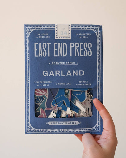 A hand holding a blue package of East End Press Garland printed paper against a plain light background. The package features white text and a window showing the colorful patterned sheets inside.