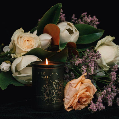A lit black candle with gold floral line art and the words 'NIGHT WALK' sits in front of a bouquet of white and peach roses, green leaves, and small purple flowers against a black background.