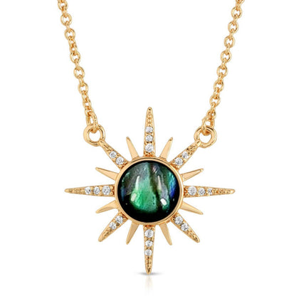 A gold sunburst pendant necklace featuring a round iridescent green labradorite stone in the center, accented with small clear pavé stones along the sun rays, shown on a white background.