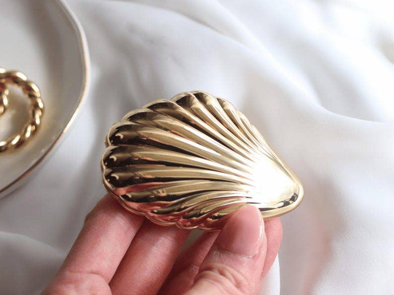 Gold shell-shaped jewelry box held by a hand on a white fabric background