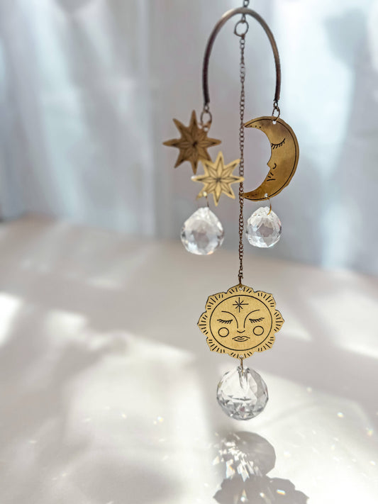 A celestial gold-toned metal suncatcher ornament with a crescent moon, stars, and a sun face charm, accented by clear faceted crystal teardrop pendants, hanging against a softly lit white background.