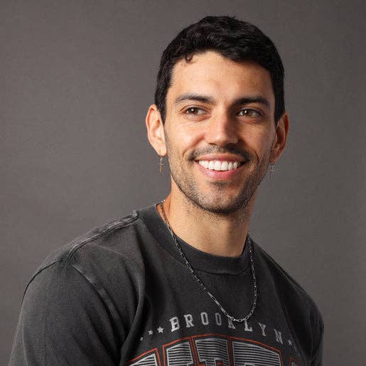 A smiling man with short dark hair and a light beard, wearing a faded black graphic t-shirt with 'BROOKLYN' printed on the front, photographed against a solid dark gray background.