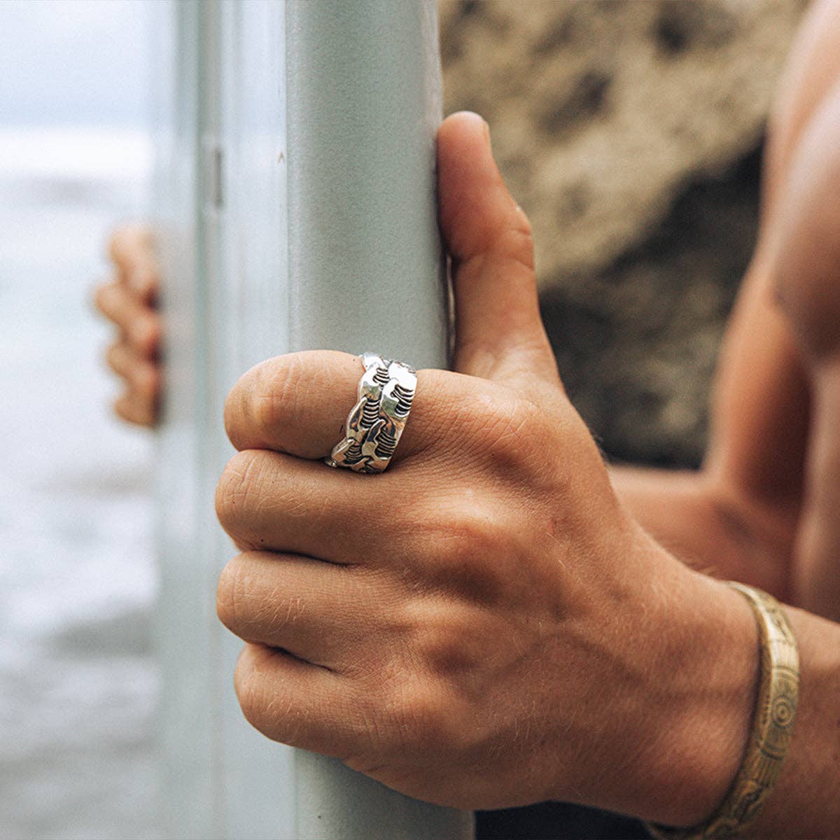 Hand holding a metal railing with a silver ring and bracelet, blurred natural background