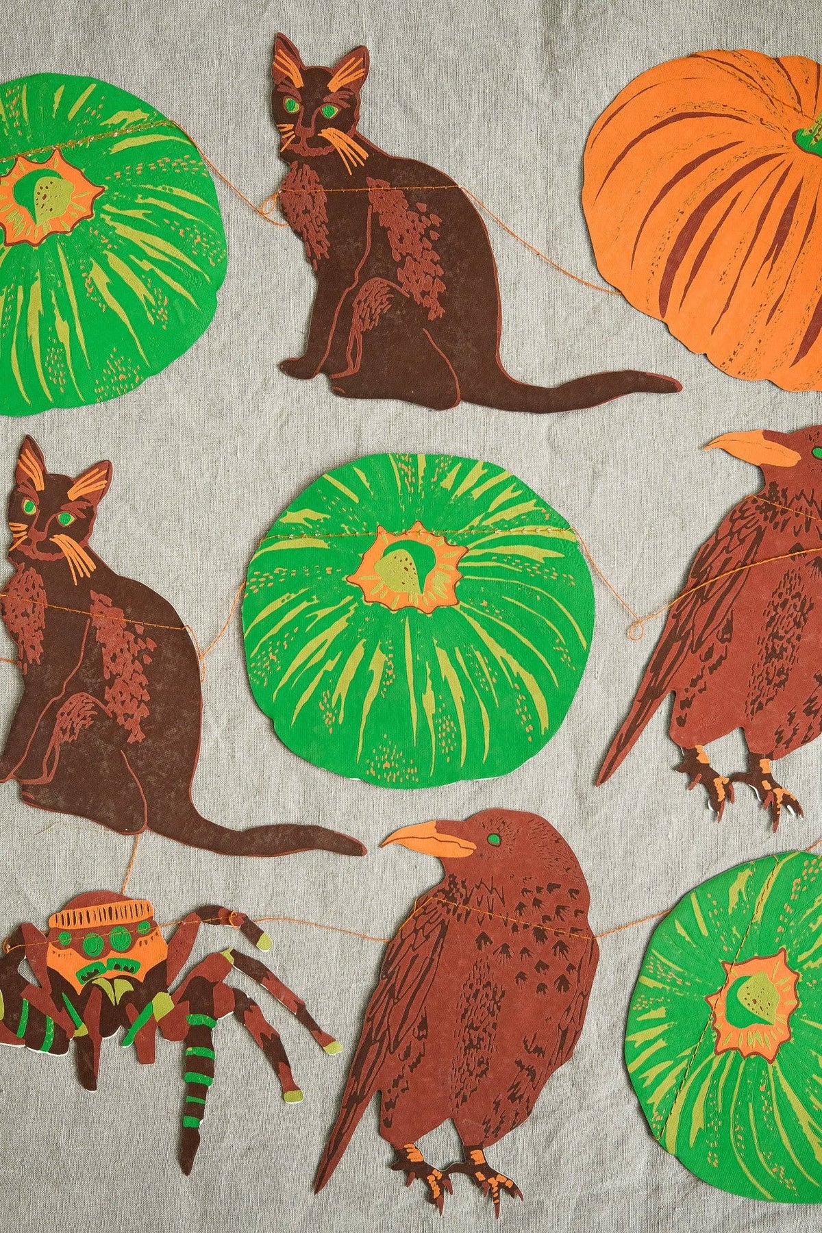 A close-up of a fabric or wallpaper pattern featuring Halloween-themed illustrations, including brown cats, orange pumpkins, green pumpkins, a brown bird, a spider, and a witch hat, all printed on a light gray textured background.