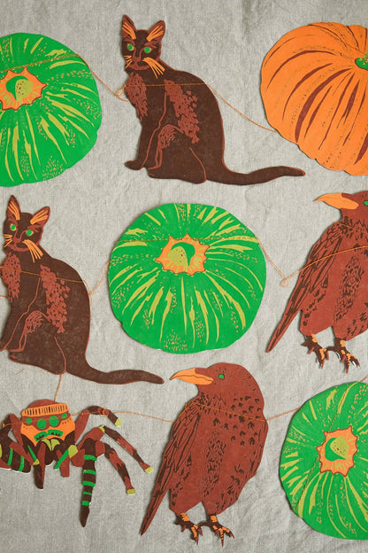 A close-up of a fabric or wallpaper pattern featuring Halloween-themed illustrations, including brown cats, orange pumpkins, green pumpkins, a brown bird, a spider, and a witch hat, all printed on a light gray textured background.