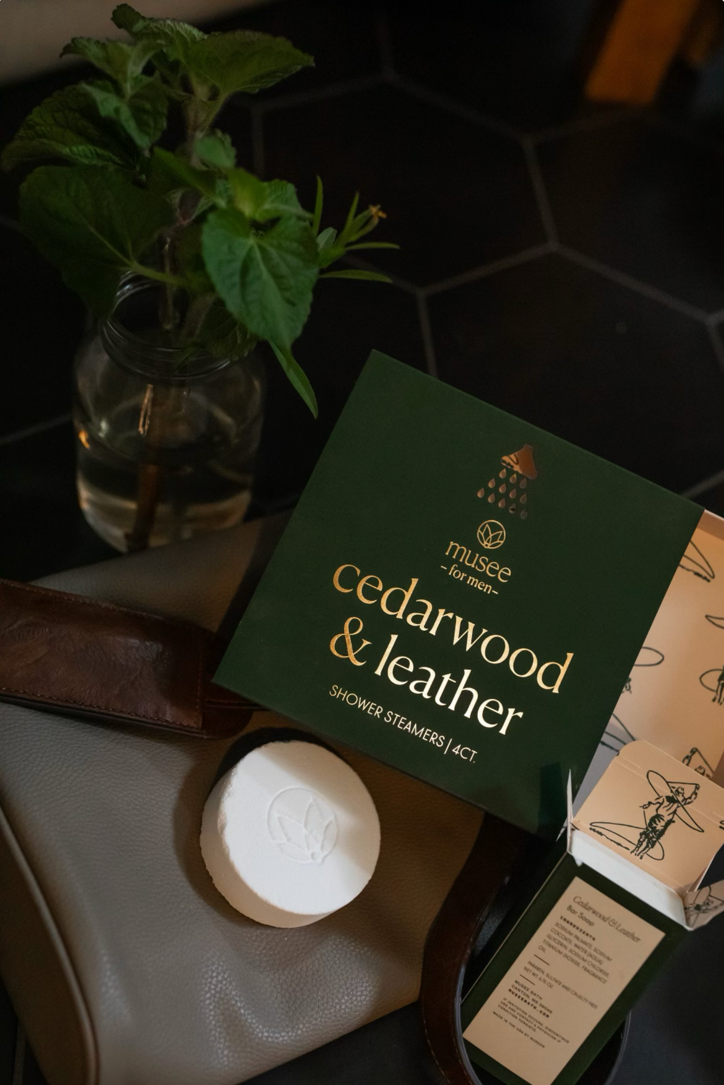 Cedarwood & Leather shower steamers by Musee on a dark surface with a plant in the background.