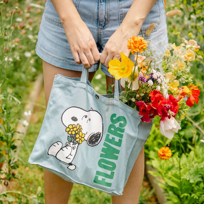 Person holding a tote bag with a cartoon character and 'FLOWERS' text, surrounded by flowers in a garden.