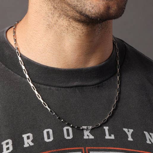 A close-up of a man's neck and lower face wearing a silver-toned metal chain necklace over a black crewneck sweatshirt.