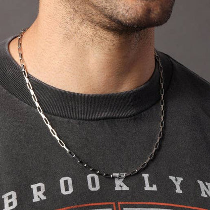 A close-up of a man's neck and lower face wearing a silver-toned metal chain necklace over a black crewneck sweatshirt.