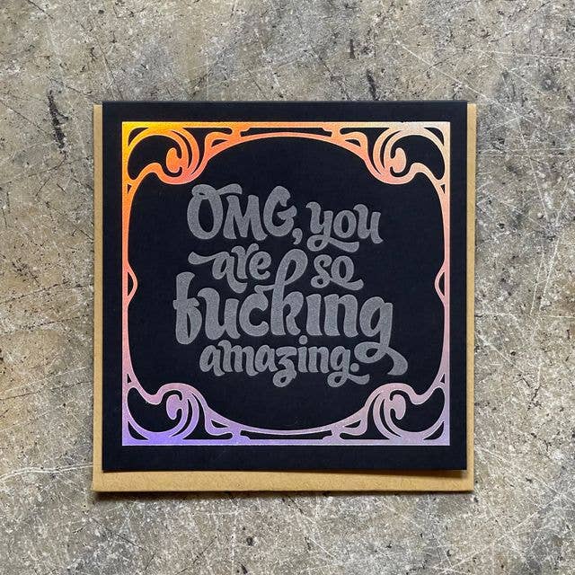 A square black greeting card with a gold border and holographic foil scrollwork, featuring the hand-lettered text 'OMG, you are so fucking amazing.' on a textured gray stone background.