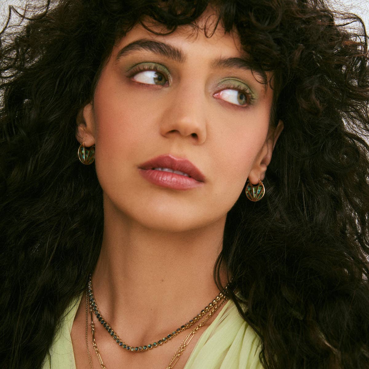 A close-up portrait of a woman with voluminous dark curly hair, wearing gold-toned layered necklaces and matching circular earrings, looking off to the side.