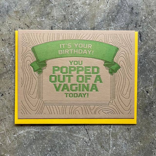 A handmade greeting card with a brown wood-grain patterned background, featuring a green banner that reads 'IT'S YOUR BIRTHDAY!' and bold green text that says 'YOU POPPED OUT OF A VAGINA TODAY!'. The card has a bright yellow border and is photographed flat on a textured gray surface.