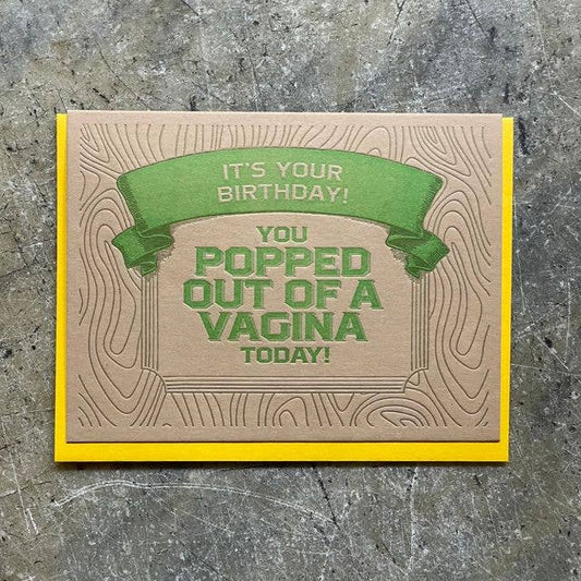 A handmade greeting card with a brown wood-grain patterned background, featuring a green banner that reads 'IT'S YOUR BIRTHDAY!' and bold green text that says 'YOU POPPED OUT OF A VAGINA TODAY!'. The card has a bright yellow border and is photographed flat on a textured gray surface.