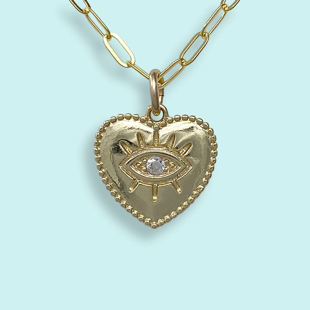 A gold heart-shaped pendant necklace featuring an eye motif with a clear central gemstone, suspended from a gold paperclip chain against a light blue background.