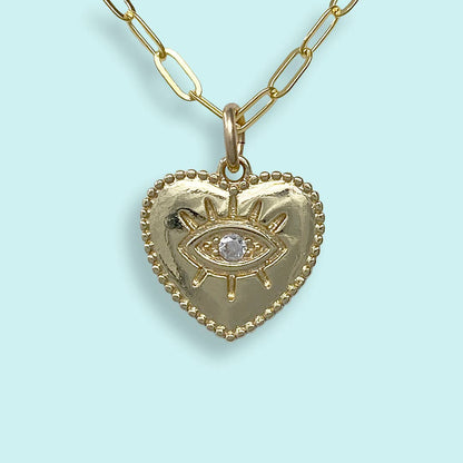 A gold heart-shaped pendant necklace featuring an eye motif with a clear central gemstone, suspended from a gold paperclip chain against a light blue background.