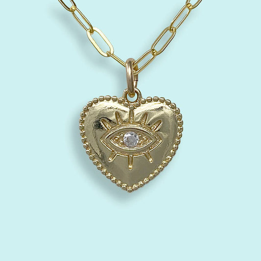 A gold heart-shaped pendant necklace featuring an eye motif with a clear central gemstone, suspended from a gold paperclip chain against a light blue background.