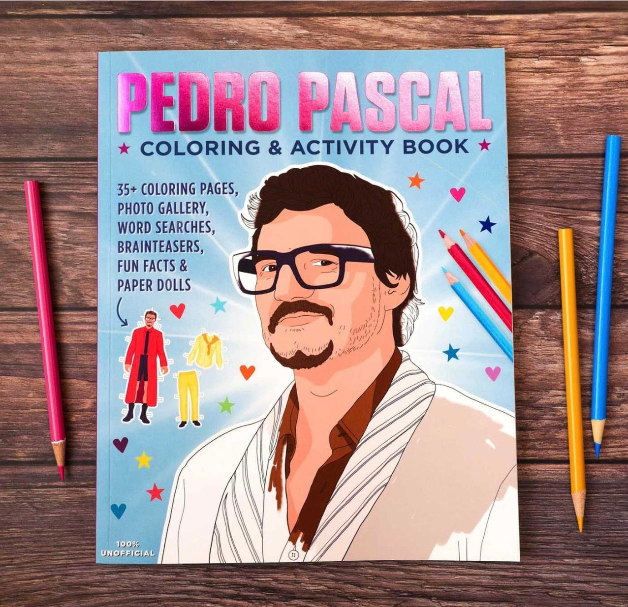 Pedro Pascal Coloring & Activity Book flat-lay on a dark wood background, flanked by colored pencils on both sides.