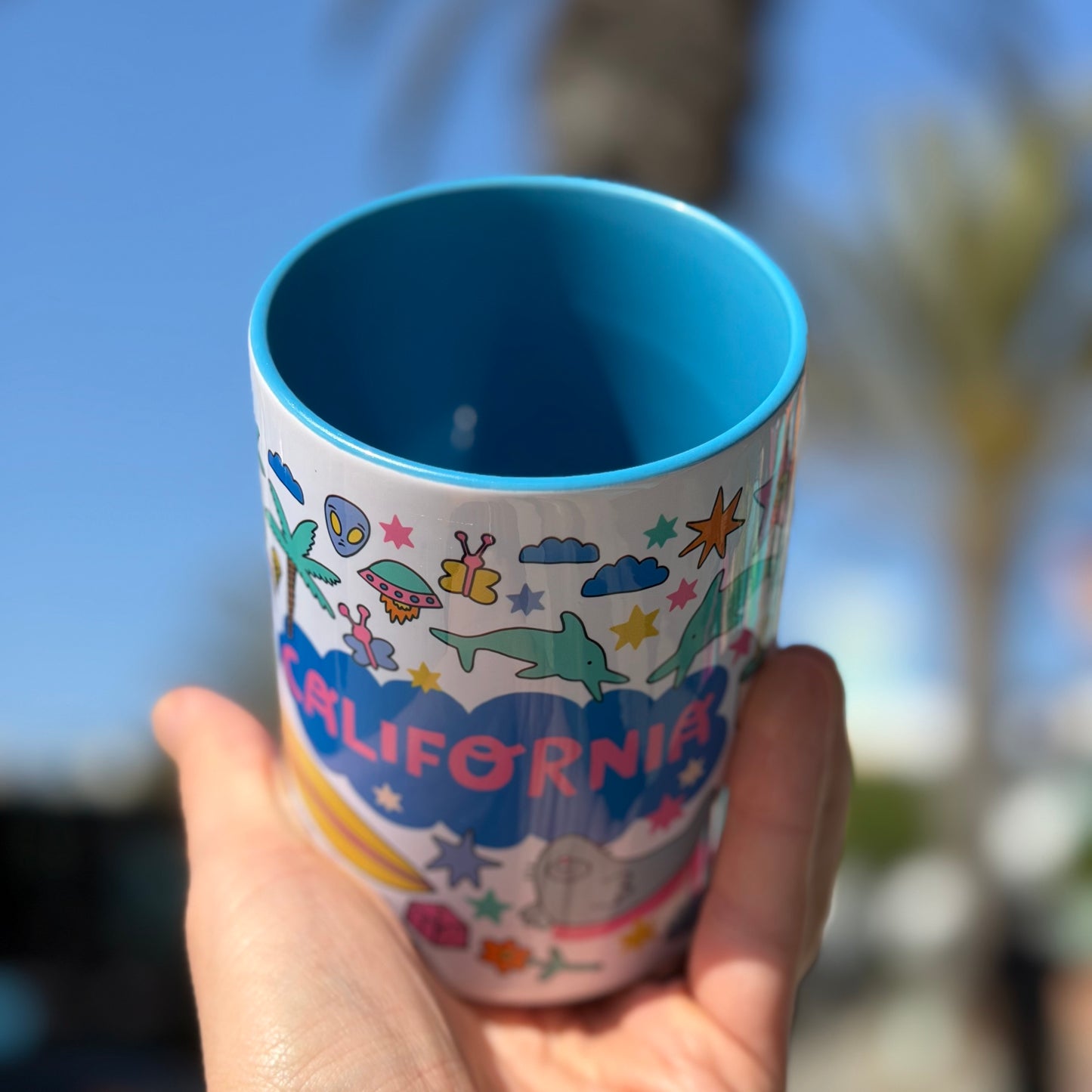 Hand holding a colorful mug with 'California' design against a blurred outdoor background