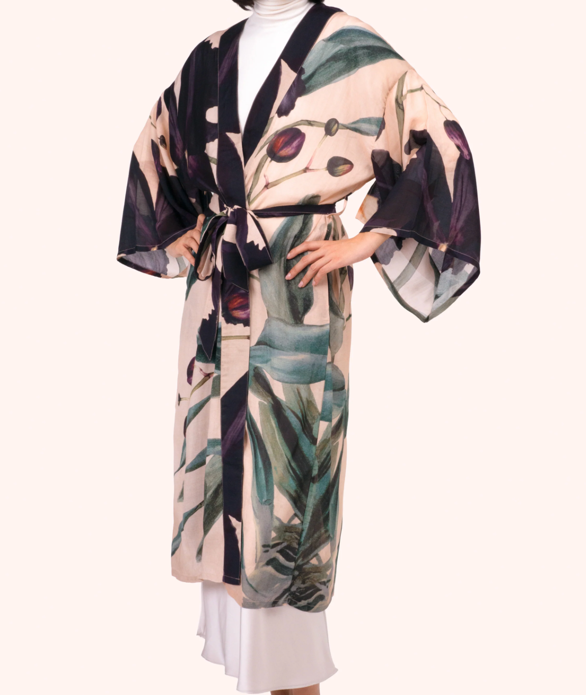 A woman wearing a kimono-style robe with a beige base and a botanical print in green, purple, and black, styled over a white hijab and white skirt against a solid light beige background.