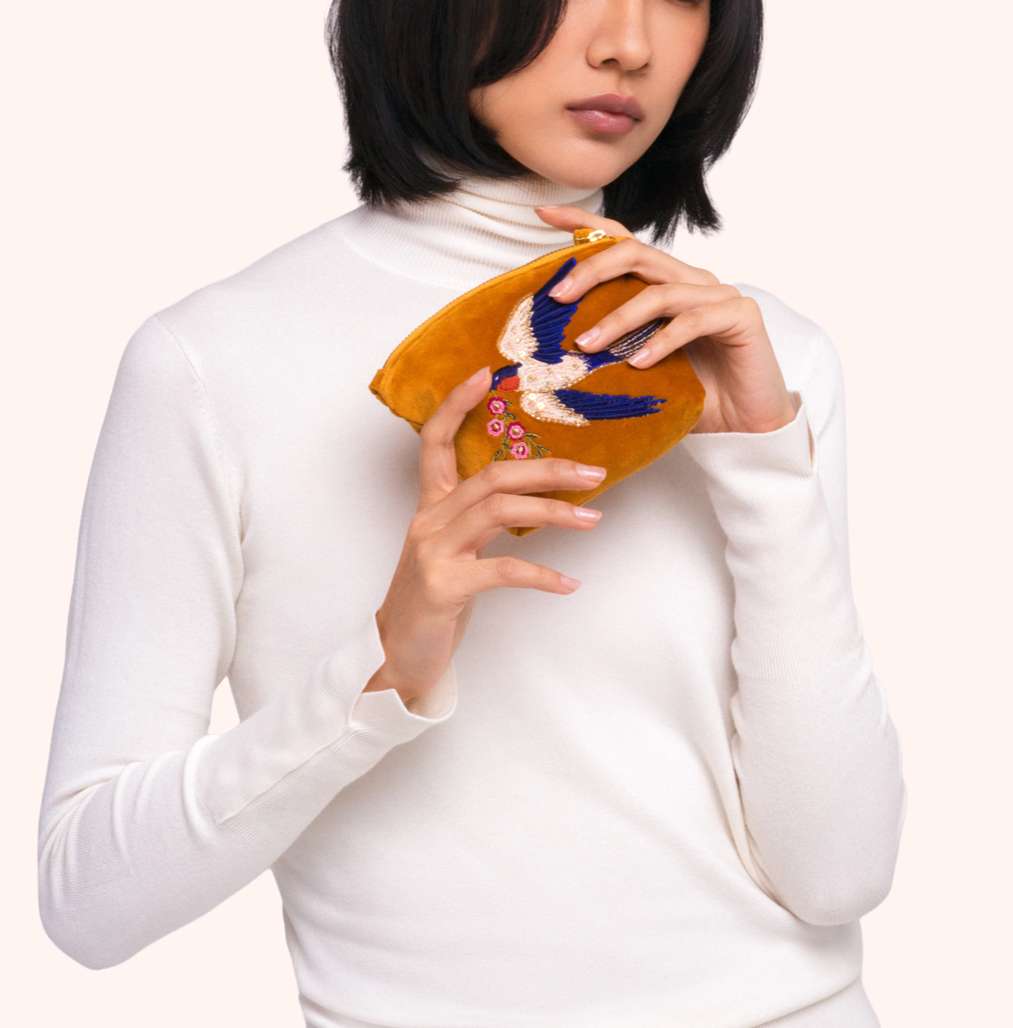 A model wearing a white turtleneck sweater holds a mustard yellow suede clutch purse decorated with a blue and white bird and colorful floral embroidery.