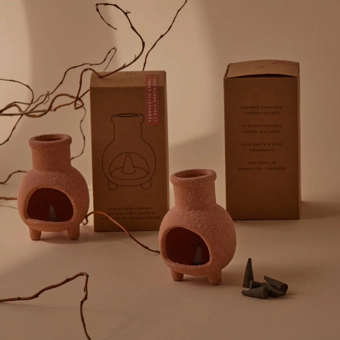 Two terracotta-colored ceramic incense burners with a textured finish, shown alongside their brown cardboard packaging boxes and a few dark incense cones on a warm beige background with decorative branches.