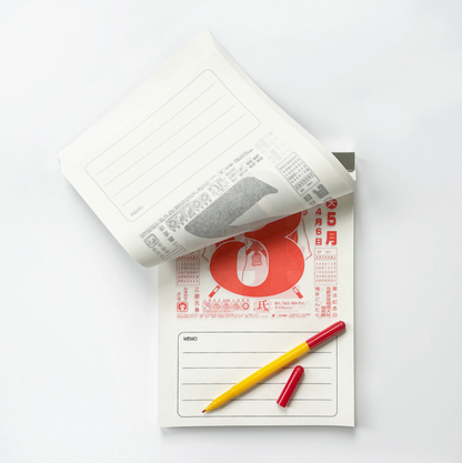 A white paper wall calendar with lined writing space is shown open, with one page flipped over. Underneath is a printed paper pad featuring a large red number 8 and Japanese text, with a yellow pen and a small red cap placed on top, all on a clean white background.