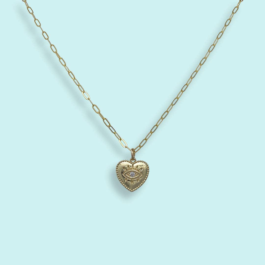 A gold heart-shaped pendant necklace featuring an eye motif with a clear central gemstone, suspended from a gold paperclip chain against a light blue background.