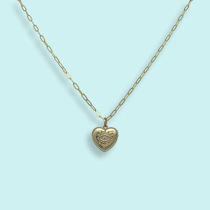 A gold heart-shaped pendant necklace featuring an eye motif with a clear central gemstone, suspended from a gold paperclip chain against a light blue background.
