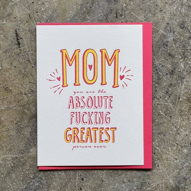 A square greeting card with a white front and bright pink border, placed on a textured gray surface. The card reads “MOM you are the ABSOLUTE FUCKING GREATEST person ever.