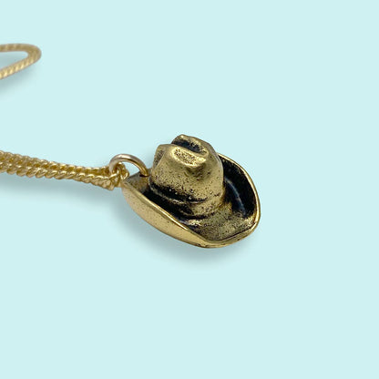 Gold cowboy hat pendant on a chain against a light blue background