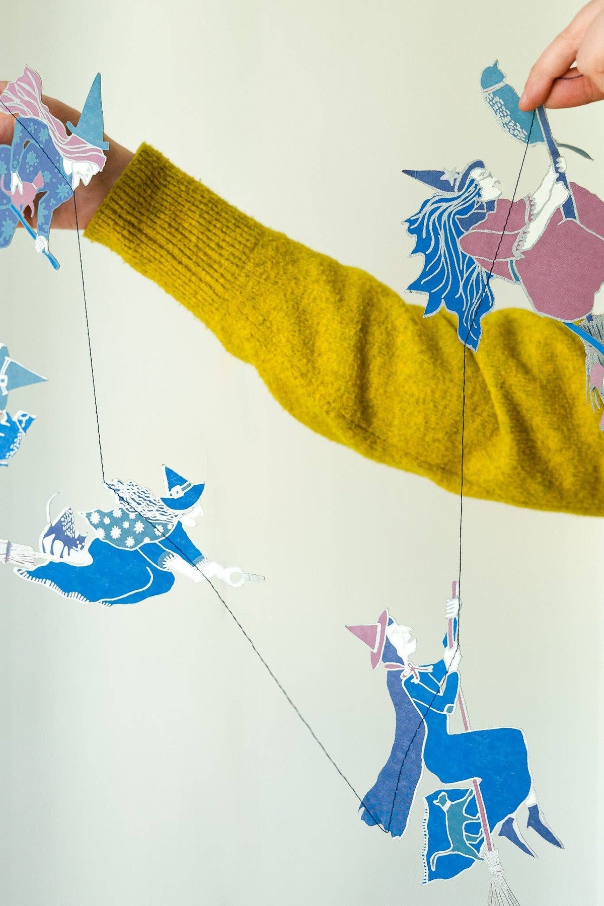 A person wearing a chunky mustard yellow knit sweater holds up a string of colorful paper cutout figures against a plain light background.