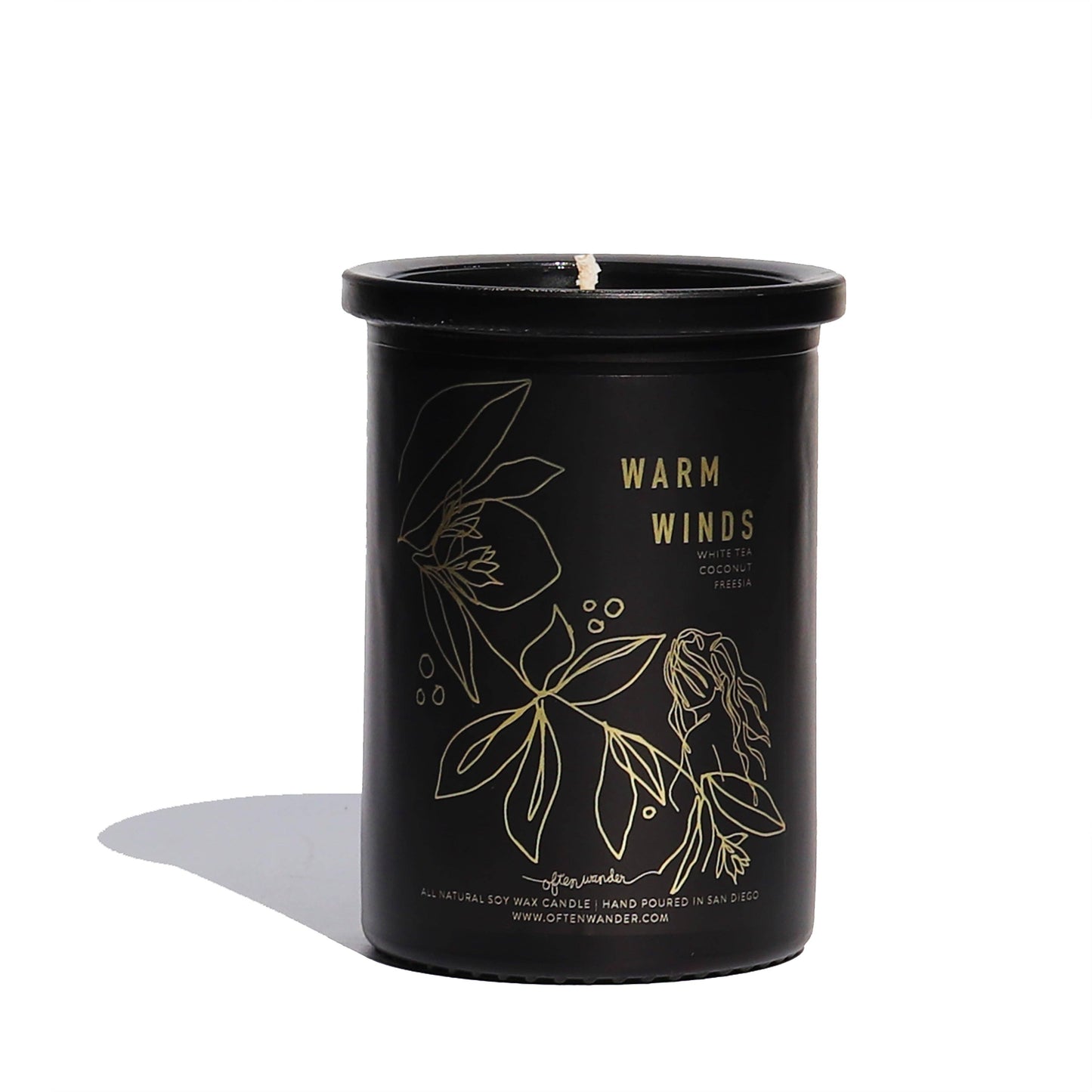A black glass jar candle with a wick at the top, featuring gold botanical line art and the text 'WARM WINDS' on the front. The candle is photographed on a clean white background with a soft shadow to the left.