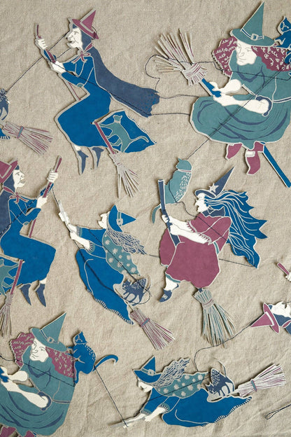 A close-up of a woven textile or rug featuring multiple stylized witch figures in blue, teal, burgundy, and cream on a beige background.
