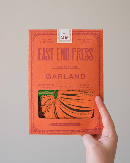 A hand holding a bright orange book titled 'EAST END PRESS GARLAND' against a plain light gray background. The cover features a window showing printed pumpkin artwork, and a small label at the top reads 'No.