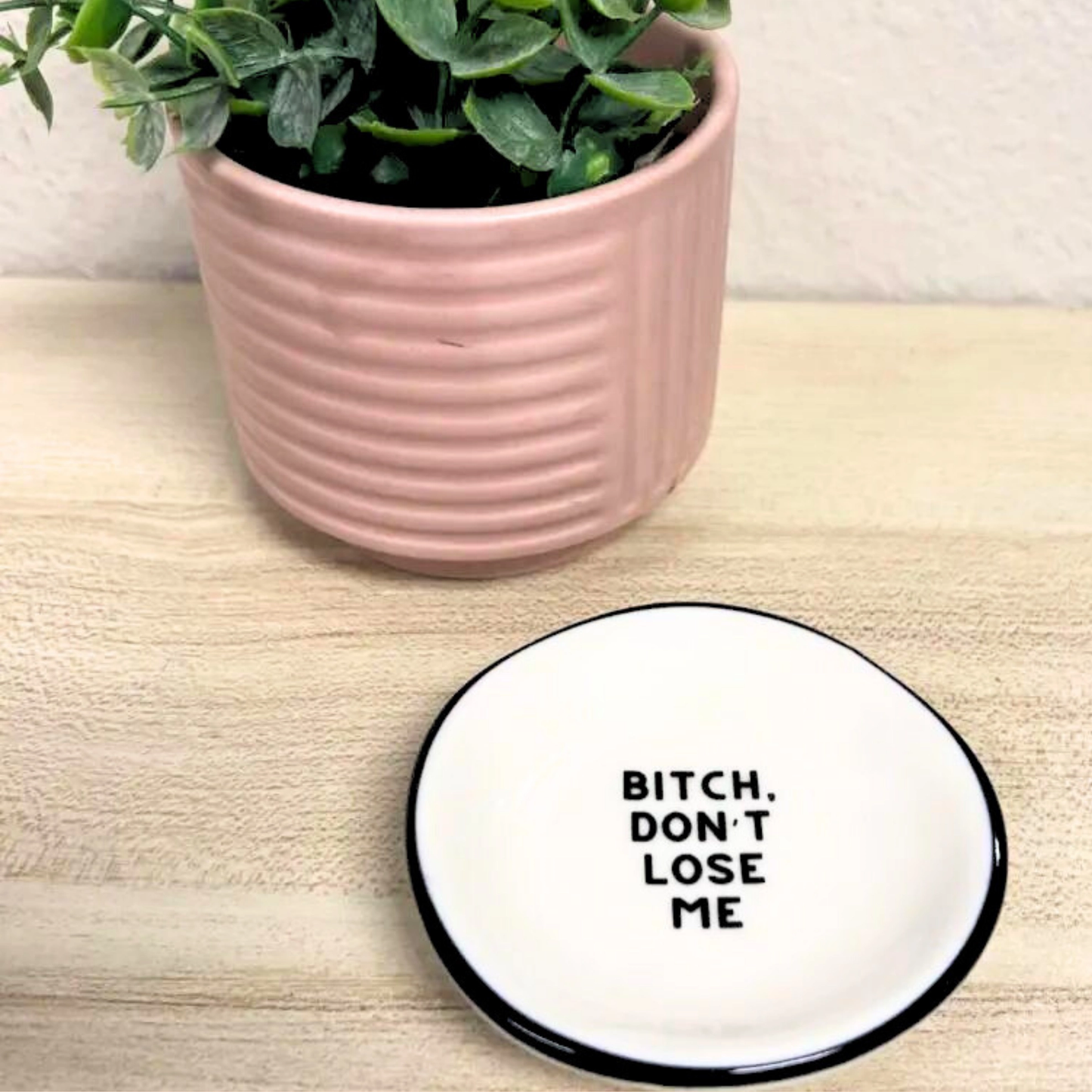 Coaster with text 'Bitch, don't lose me' next to a pink pot with a plant on a wooden surface