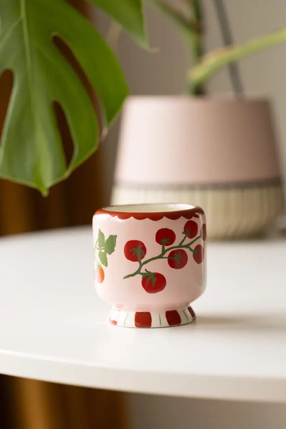 Decorative ceramic candle vase with tomato vine design on a white surface, with a blurred plant and lamp in the background.