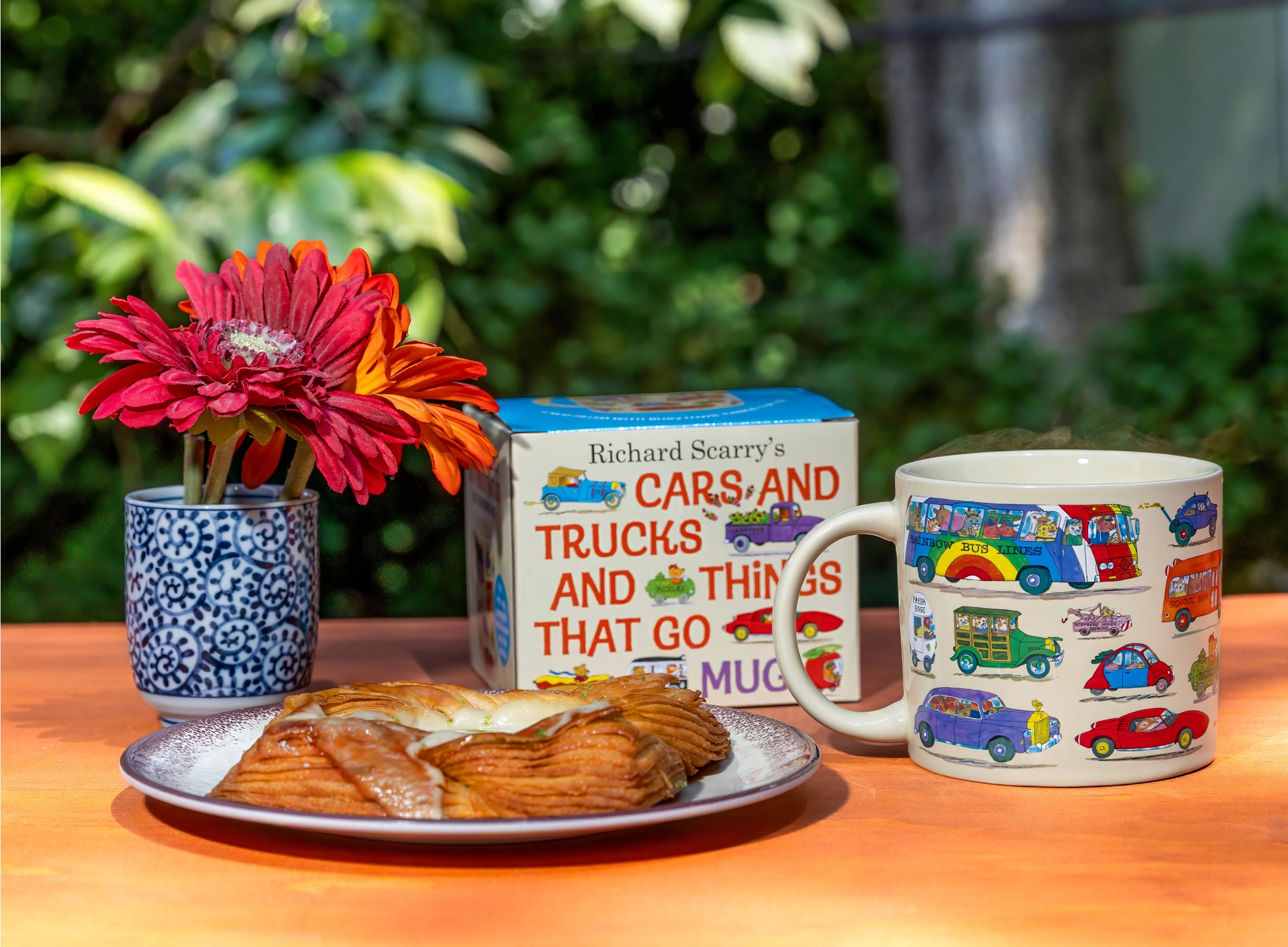 Cup with car designs, cookies, a mug with flowers, and a book titled 'Cars and Trucks and Things That Go' on a table outdoors.