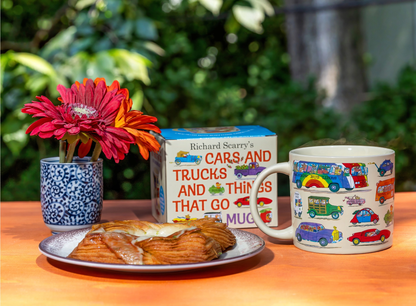 Cup with car designs, cookies, a mug with flowers, and a book titled 'Cars and Trucks and Things That Go' on a table outdoors.