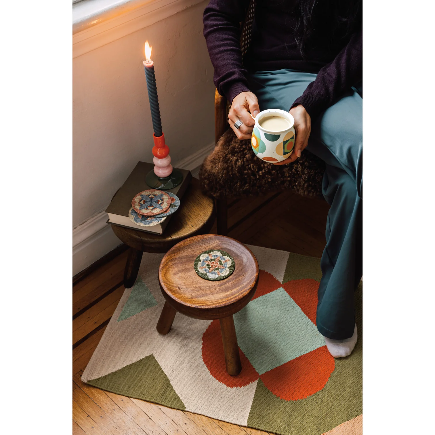 Person sitting on a wooden stool holding a mug with a candle and small table nearby.